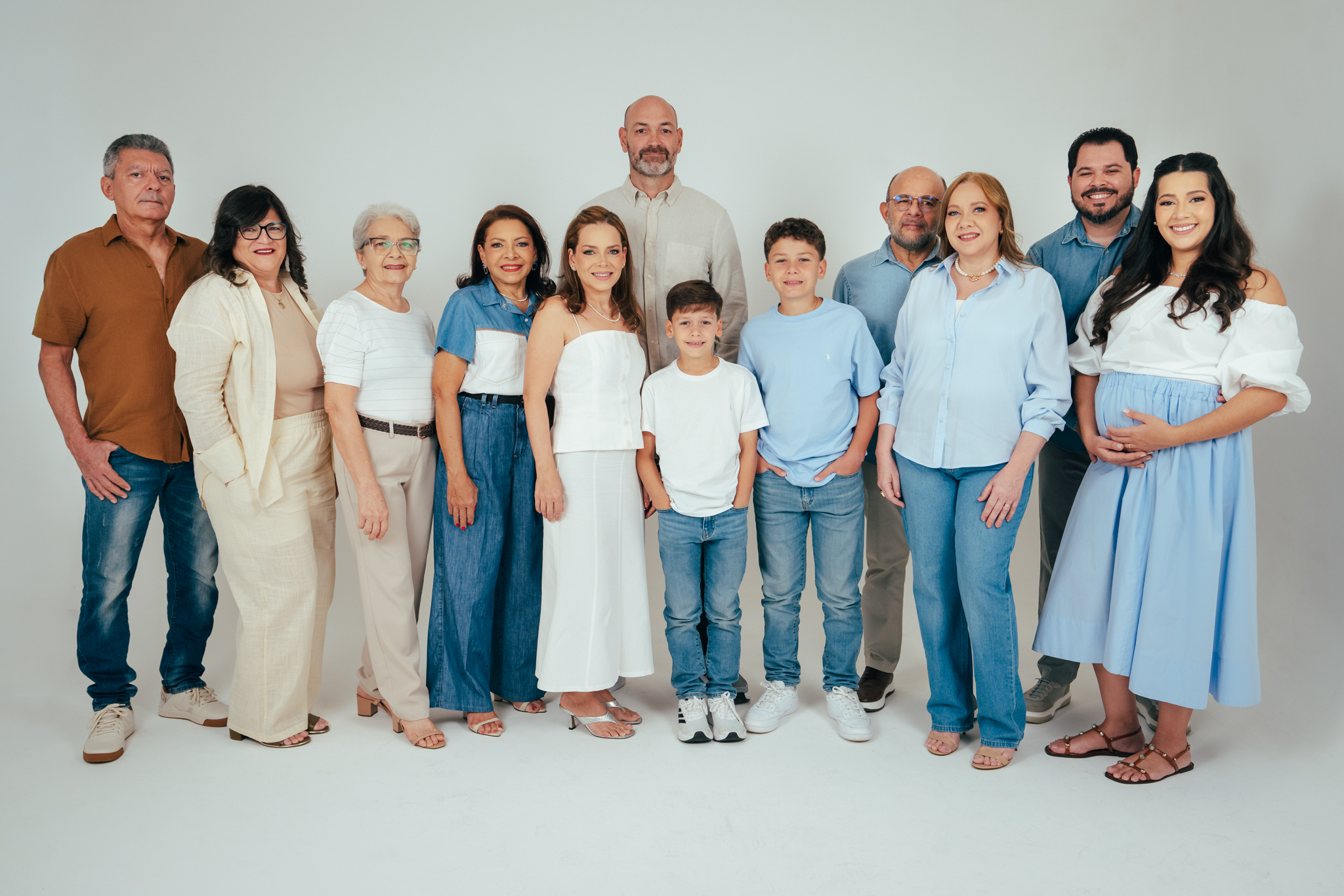 Retrato de família posada em estúdio com fundo branco, mostrando onze pessoas de idades variadas vestidas em tons neutros e azuis.