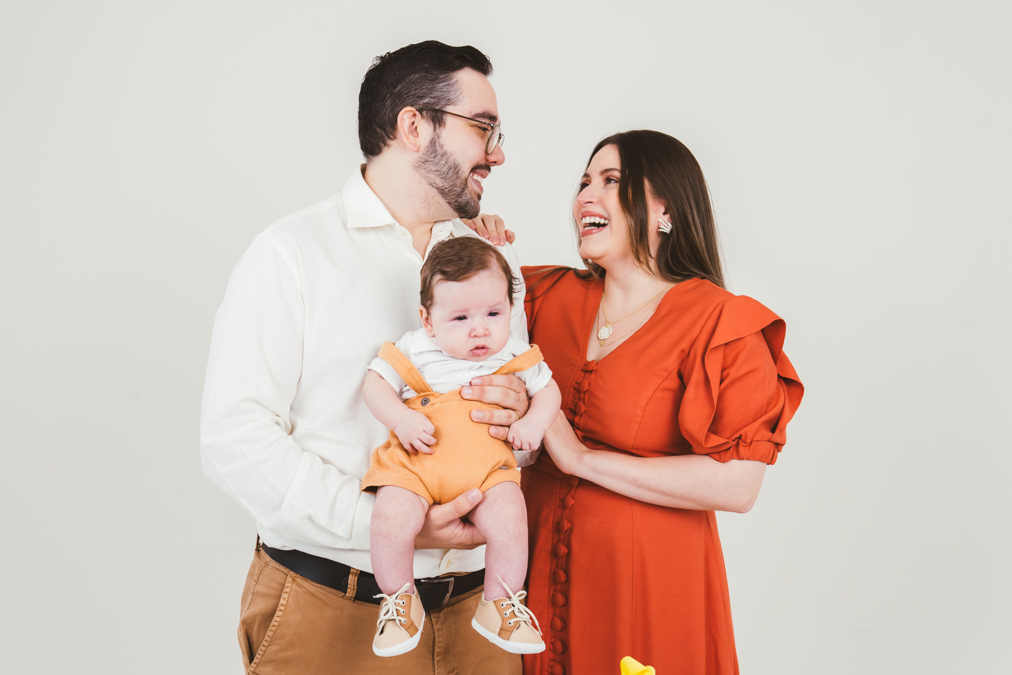 Homem com camisa branca e mulher de vestido laranja sorriem enquanto seguram um bebê com jardineira mostarda diante de fundo neutro.