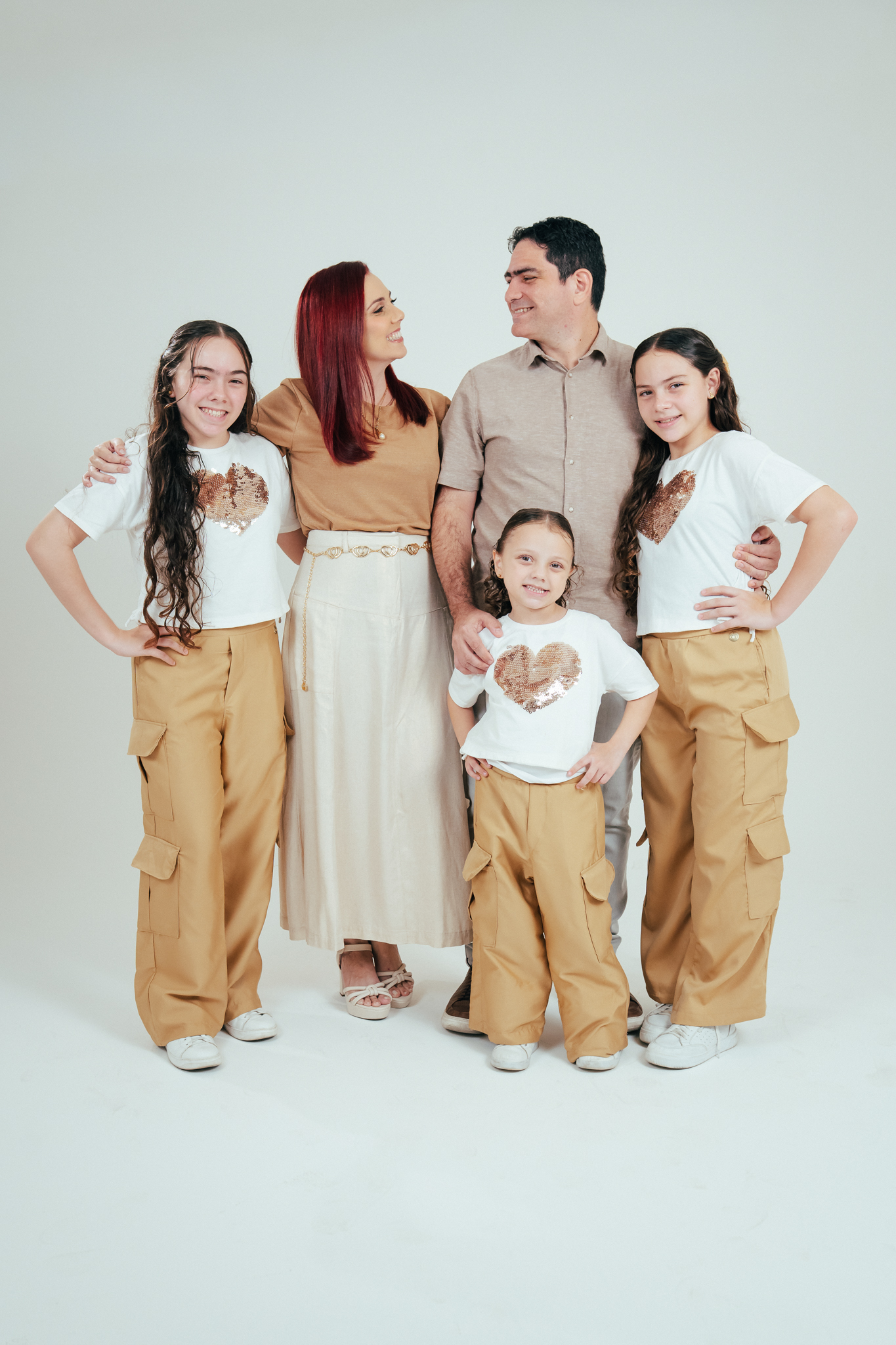 Família com cinco pessoas posa em fundo claro, usando roupas em tons bege e branco, com as filhas vestindo camisetas com coração de lantejoulas.