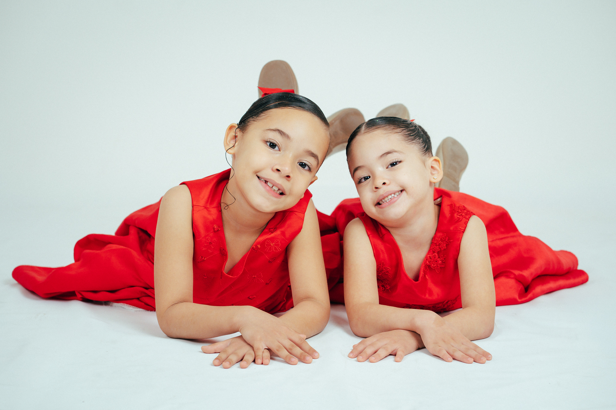 Duas meninas sorridentes de vestidos vermelhos deitadas de bruços em um fundo branco, apoiadas nos cotovelos.
