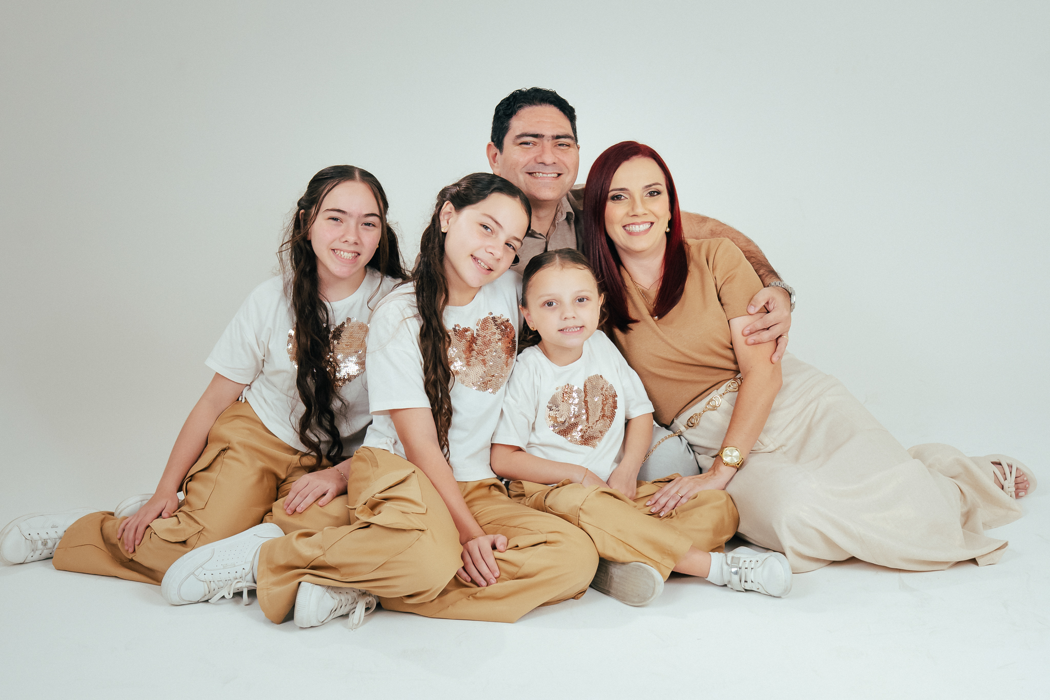 Família sorri abraçada no estúdio, vestindo blusas brancas com lantejoulas e calças em tons de bege.