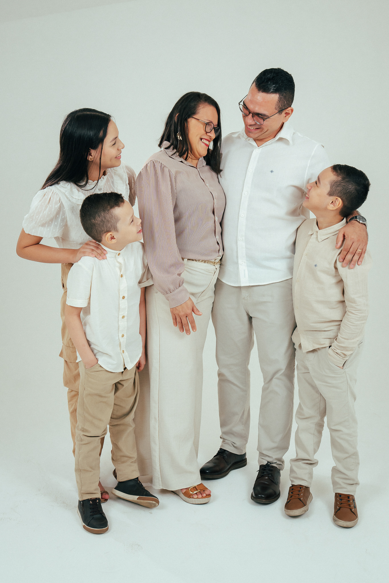 Família de cinco pessoas posa sorridente em estúdio com fundo branco, vestindo roupas em tons claros.