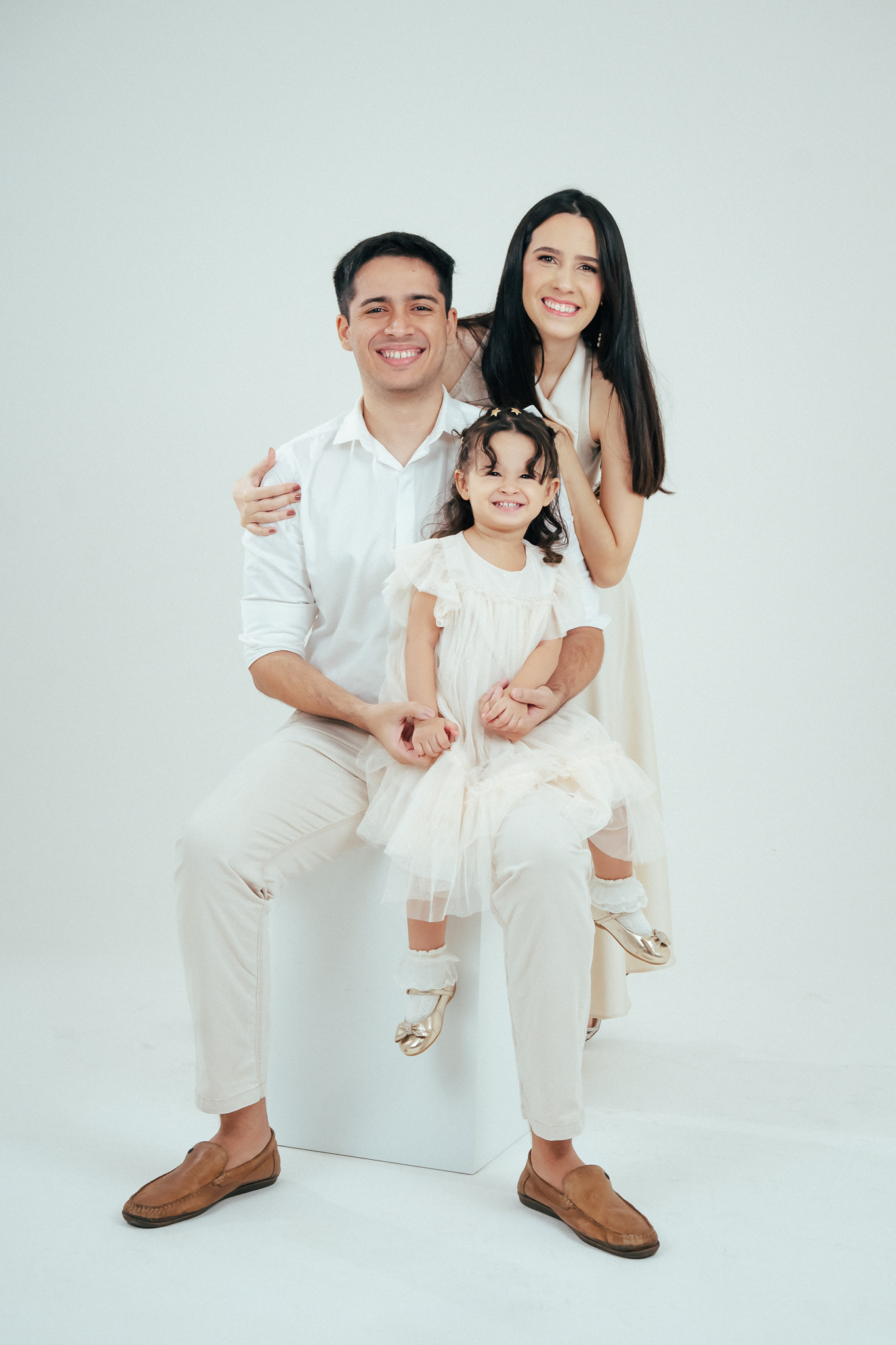 Família sorri em estúdio com fundo branco; pai sentado segura a filha pequena e mãe está em pé logo atrás, todos vestidos com roupas claras.