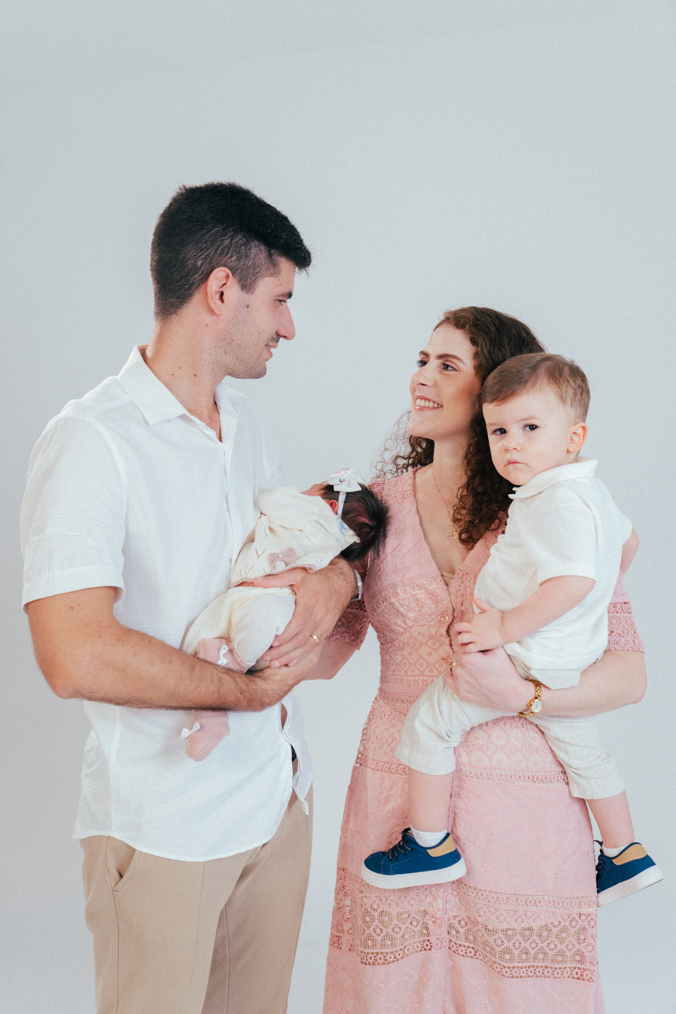 Pai e mãe sorriem, segurando um bebê recém-nascido e um menino menor, contra fundo branco.