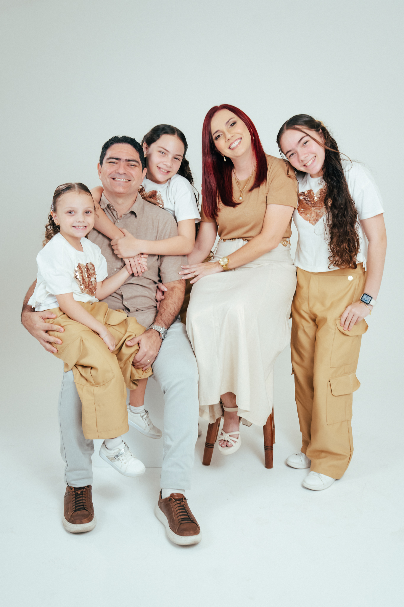 Família sorri reunida em estúdio: pai sentado com a caçula no colo, mãe sentada ao lado e duas filhas em pé, todos com roupas em tons bege e branco.
