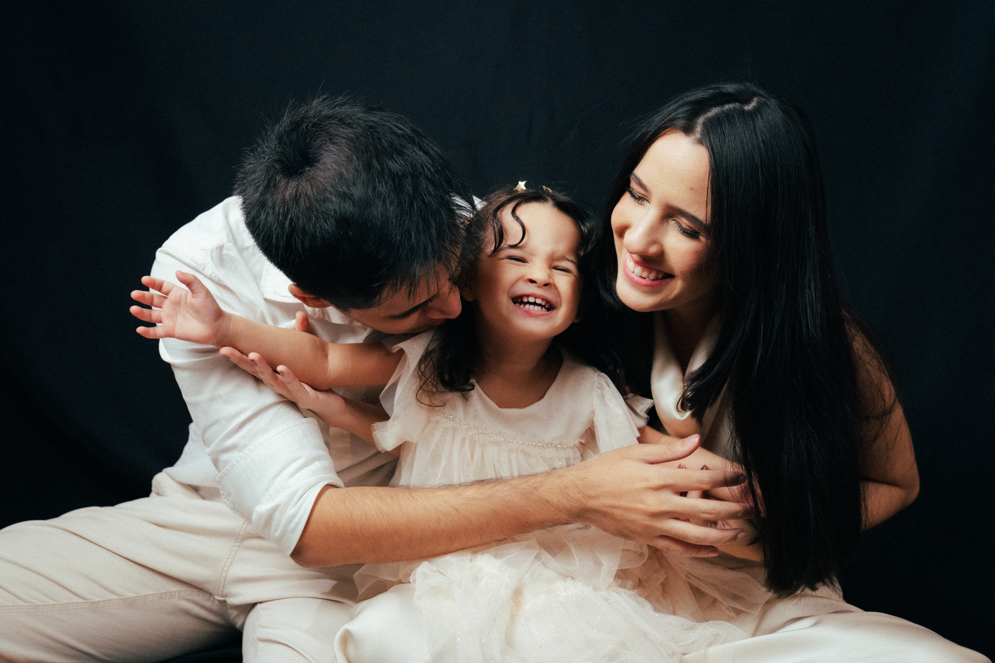 Pai, mãe e filha sorridentes vestidos com roupas claras em um estúdio com fundo preto.
