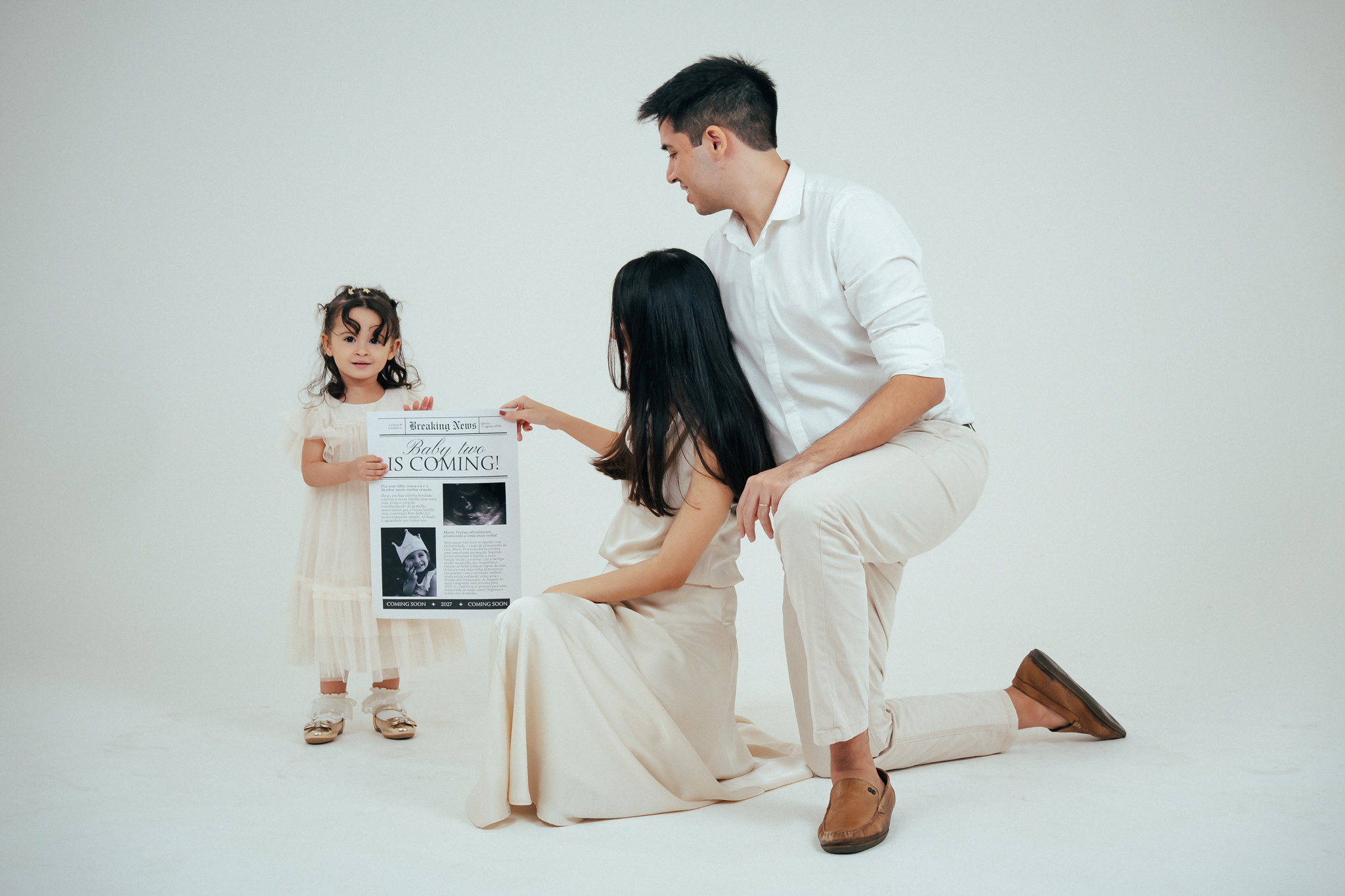 Família sorri em fundo branco enquanto segura um cartaz que anuncia a chegada de um novo bebê.