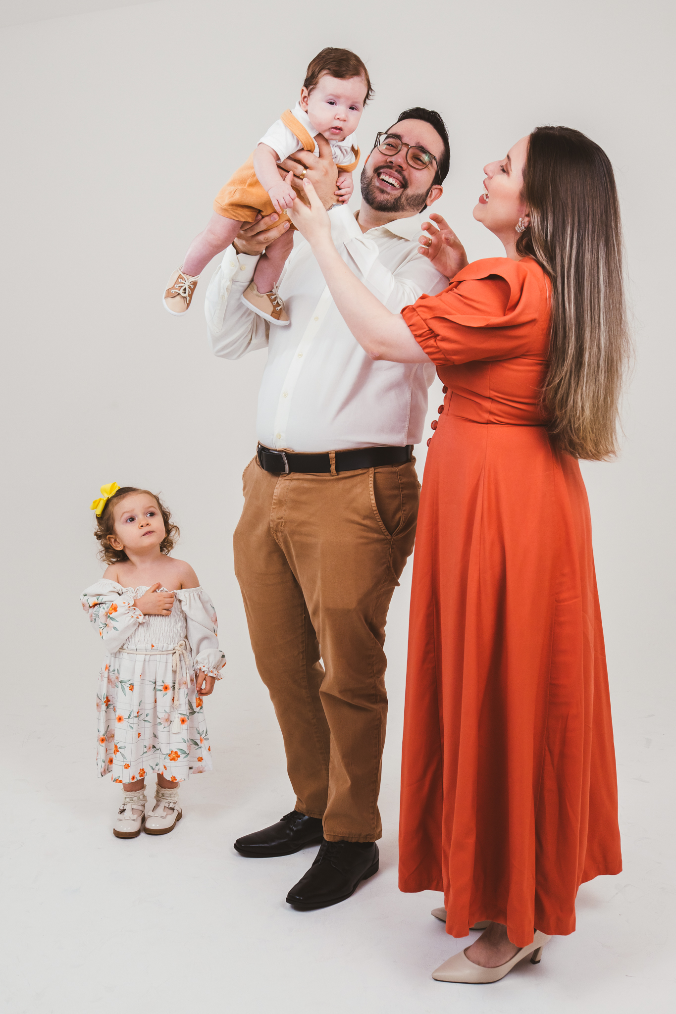 Família sorri reunida no estúdio; o pai levanta o bebê no alto, a mãe de vestido laranja olha para cima e a menina observa ao lado.