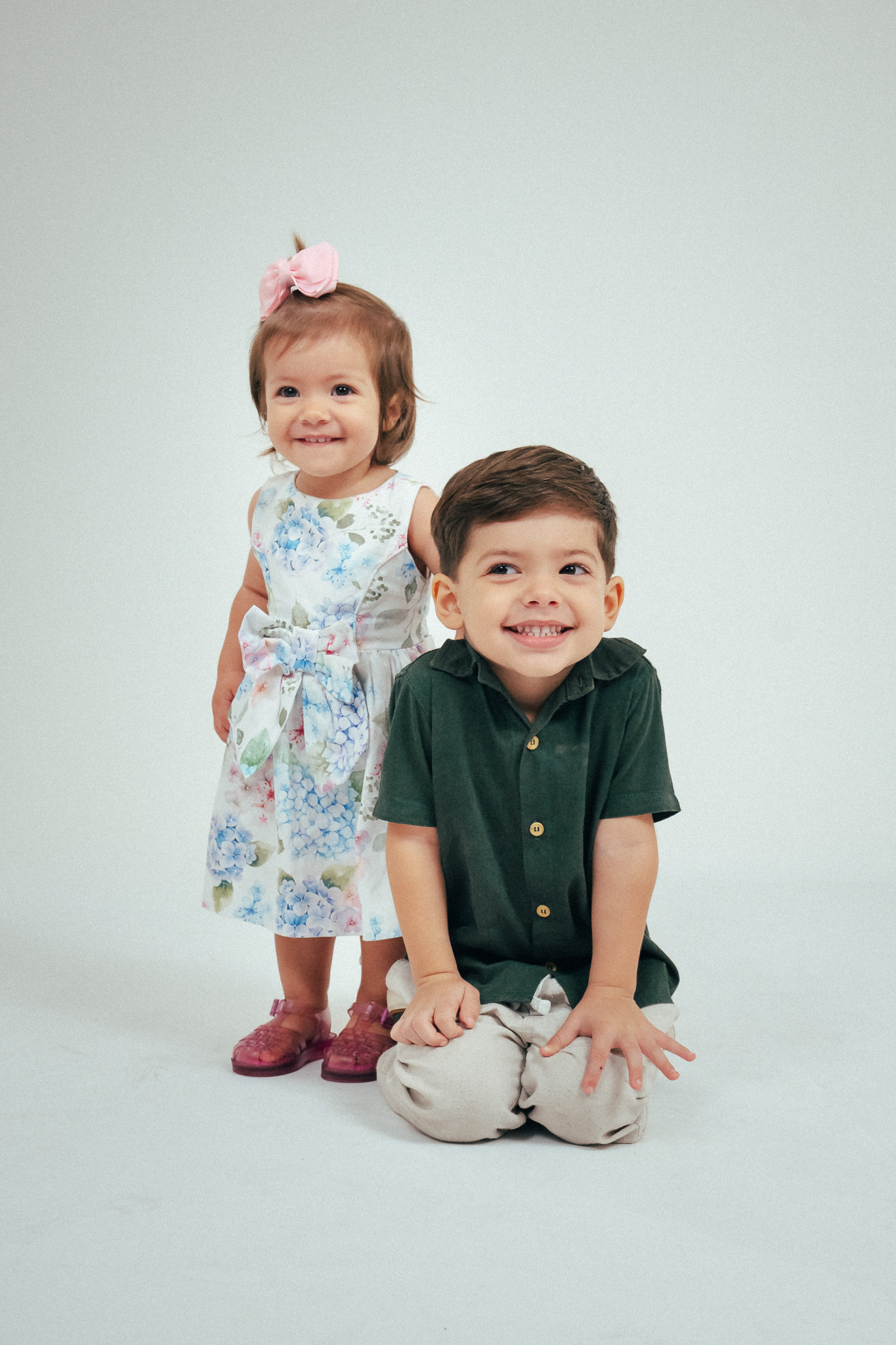 Menina em pé de vestido floral com laço e menino ajoelhado de camisa verde sorriem juntos diante de fundo branco.