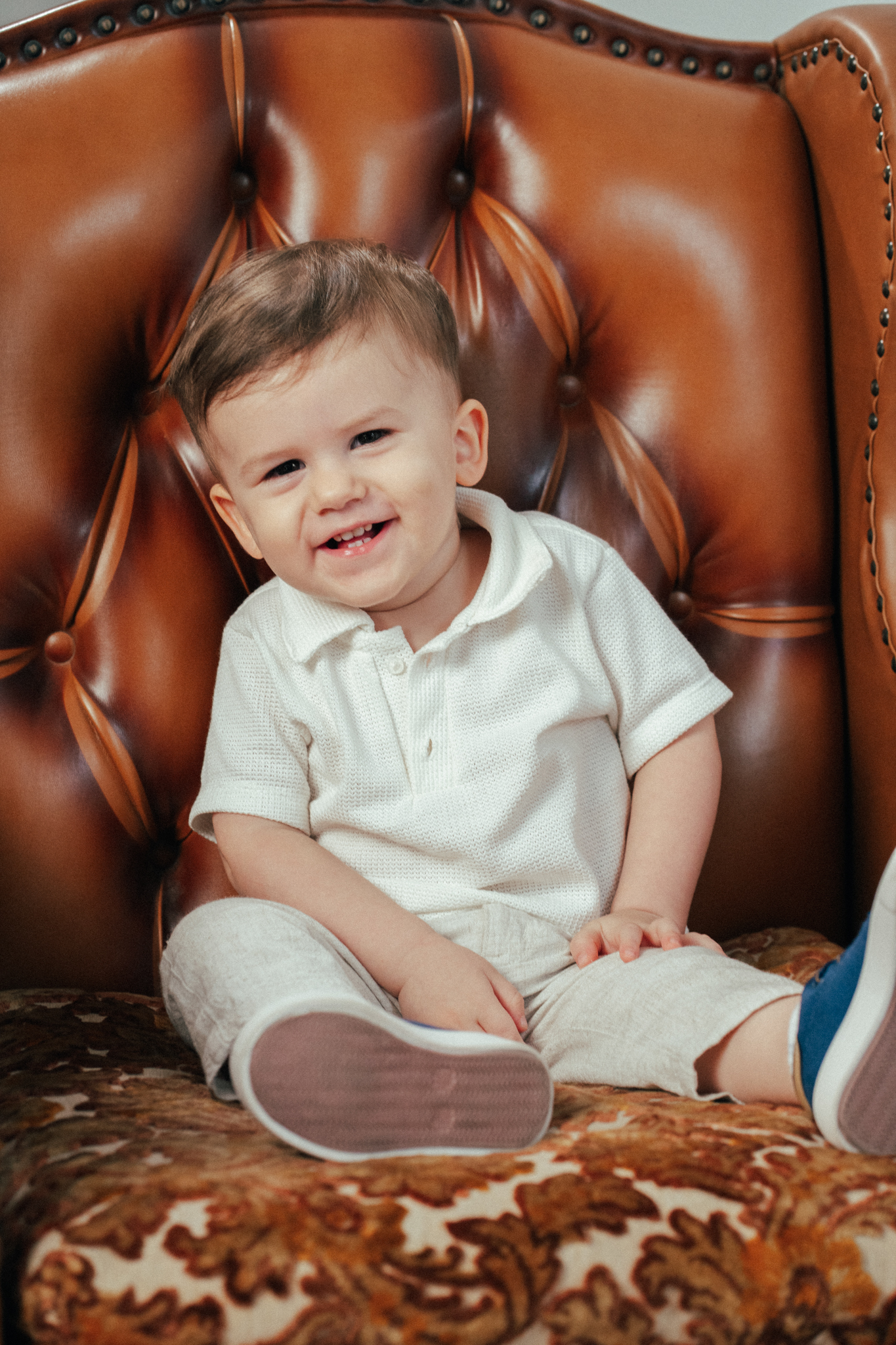 Menino sorridente de camisa branca e calça clara sentado em uma poltrona de couro marrom.