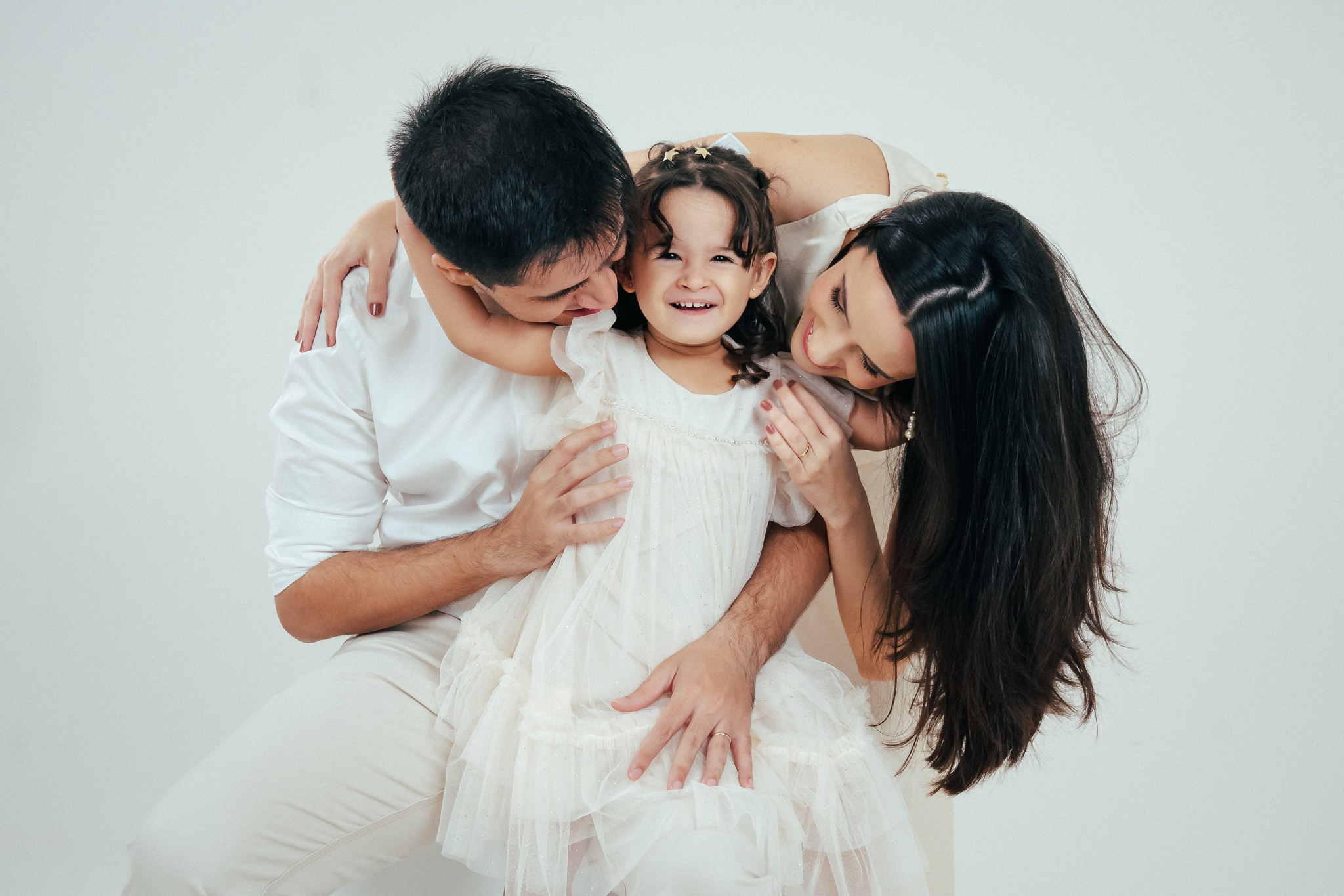 Pai, mãe e filha sorridentes vestidos de branco posam abraçados em um fundo neutro de estúdio.