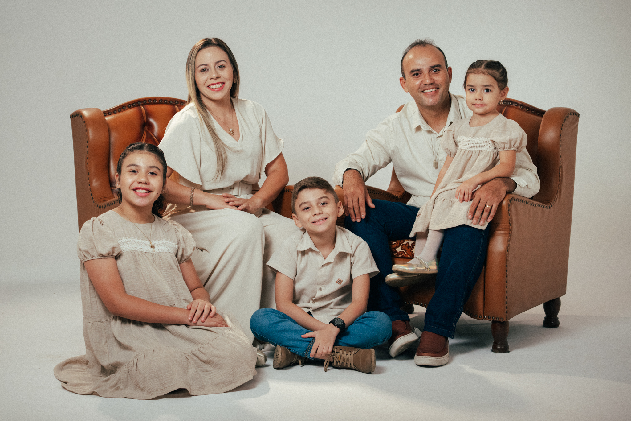 Família sorri em estúdio: casal está sentado em poltrona marrom com duas filhas e um filho ao redor.