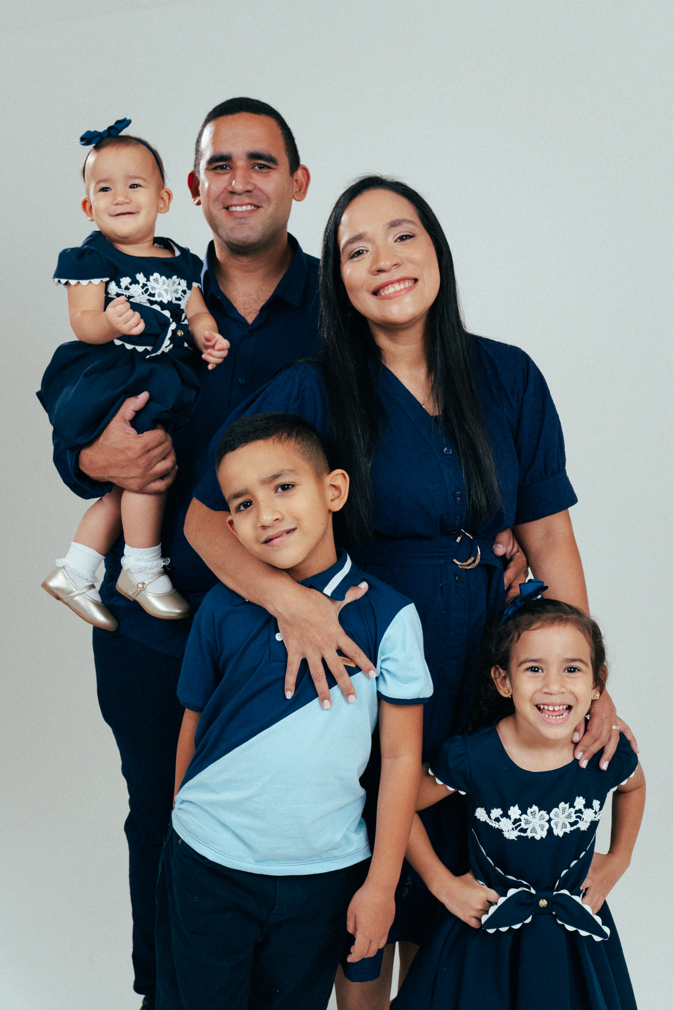 Família sorri unida vestindo azul em estúdio; pai segura bebê, mãe abraça filho e filha sorri ao lado.