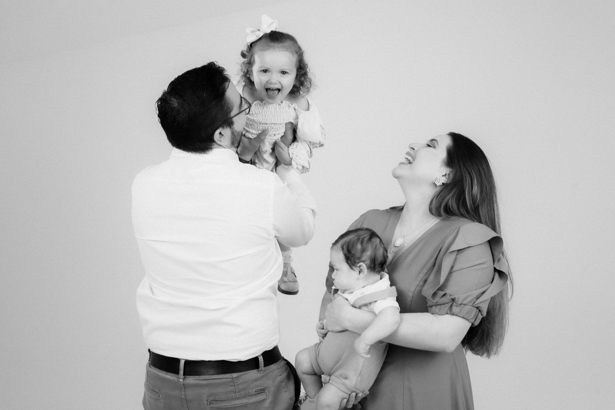 Foto em preto e branco de uma família sorridente em estúdio: o pai ergue uma menina no alto, enquanto a mãe segura um bebê no colo.