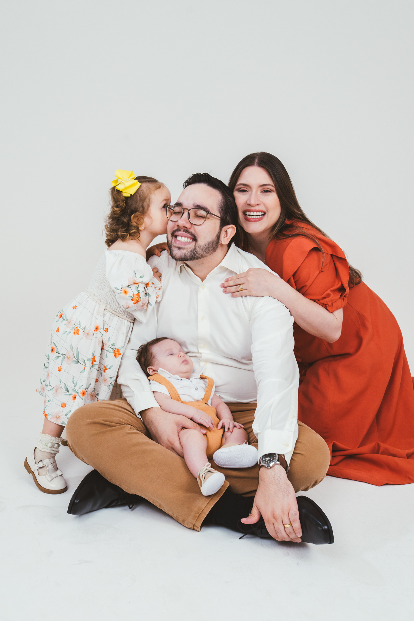 Família posa sorrindo em estúdio com fundo branco, com a filha beijando o pai e o bebê dormindo no colo dele.