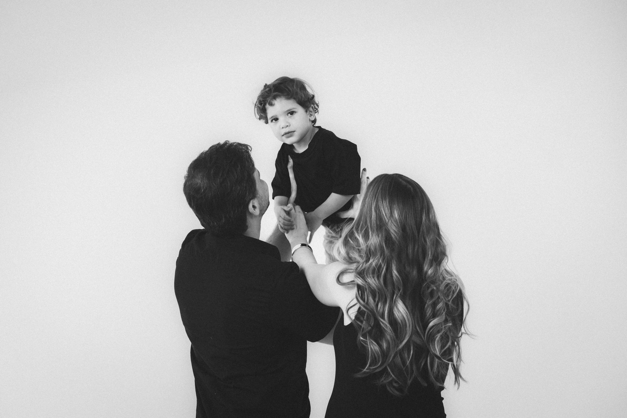 Foto em preto e branco de pai e mãe erguendo no alto um menino pequeno, vestidos com roupas pretas, contra fundo branco e neutro.