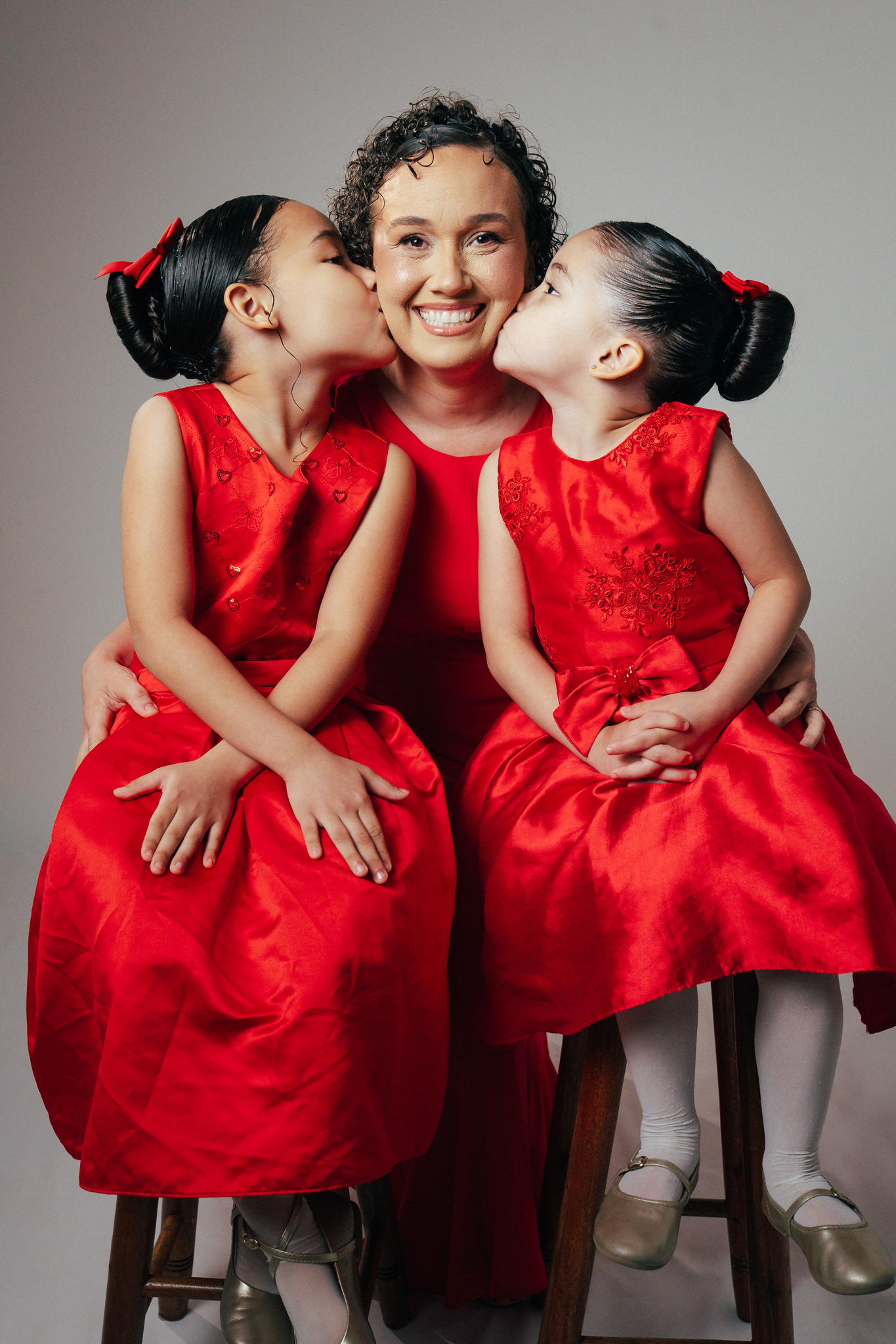 Mãe sorri no centro enquanto duas meninas com vestidos vermelhos idênticos beijam suas bochechas.