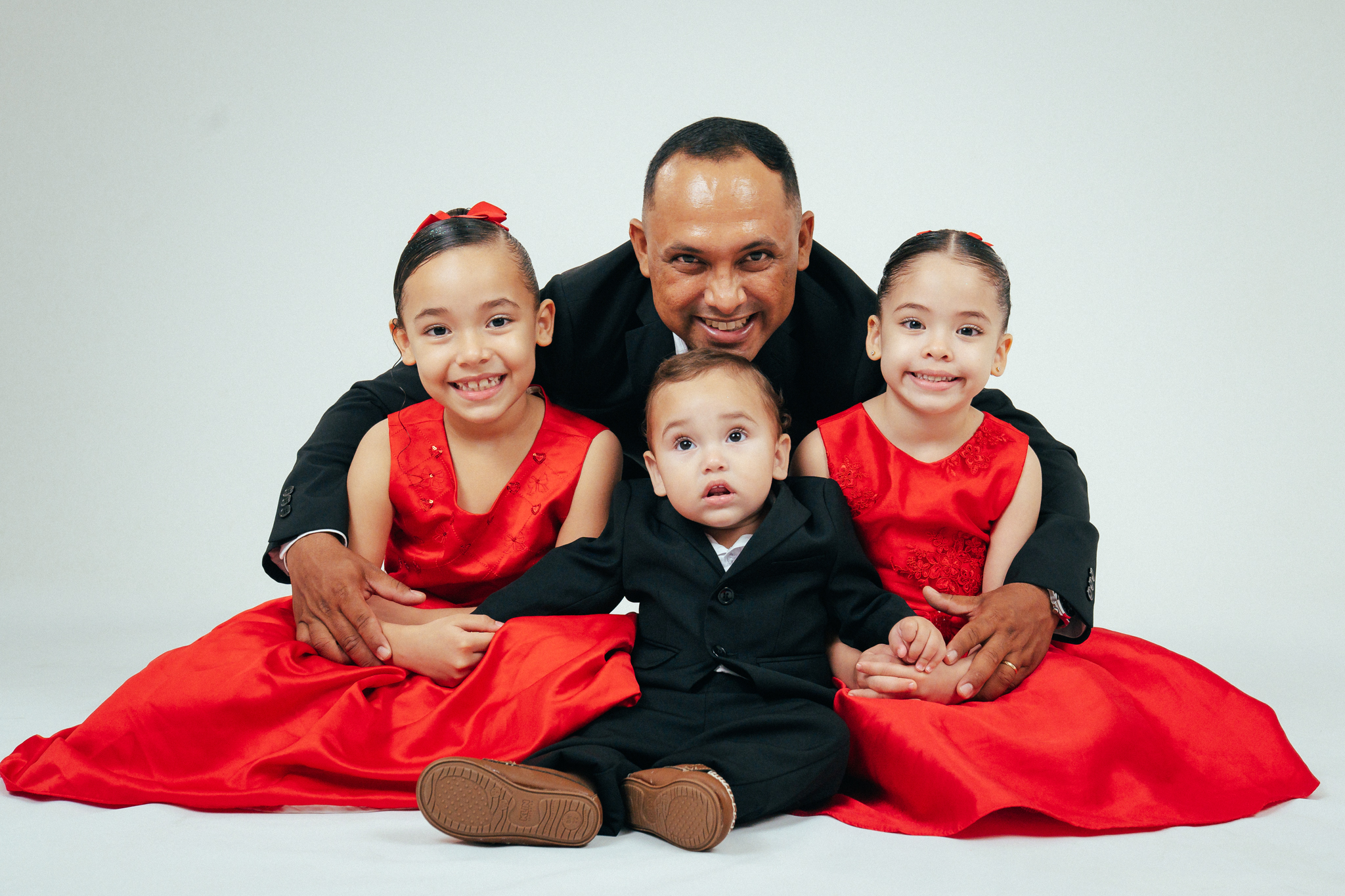 Pai sorri abraçado a duas meninas de vestido vermelho e um bebê de terno preto em estúdio.