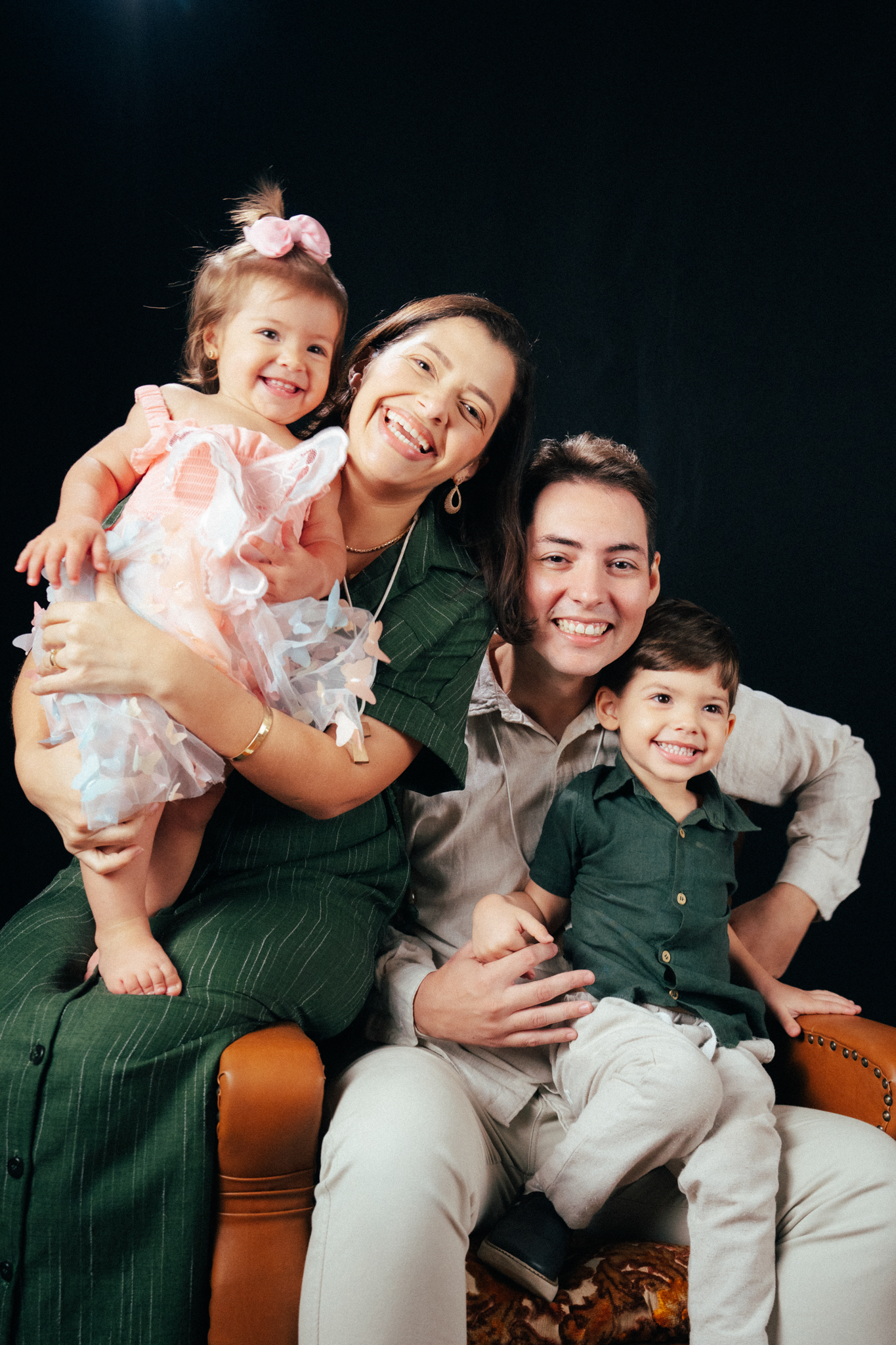 Família sorri em poltrona: mulher segura bebê de vestido rosa e homem segura menino de camisa verde em fundo preto.
