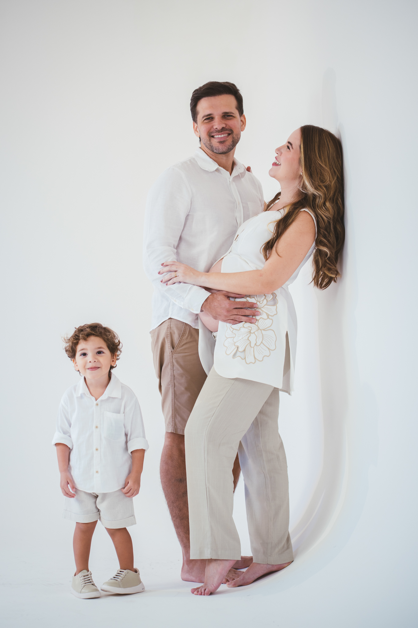 Homem e gestante sorriem abraçados junto à parede branca, enquanto um menino em pé posa à frente deles, todos vestidos em tons claros!
