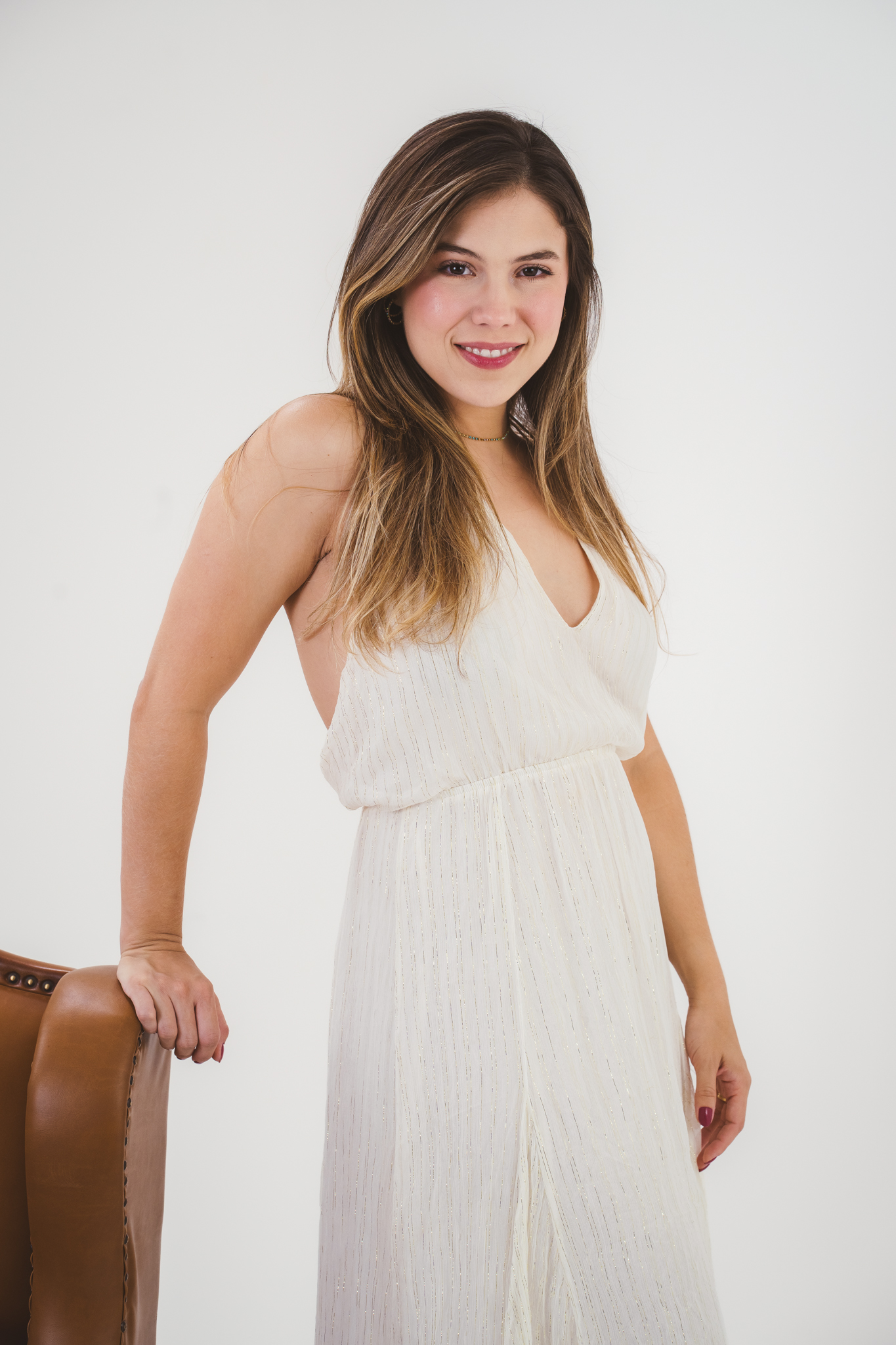 Retrato de mulher sorridente com vestido branco longo, apoiada em poltrona de couro marrom, em fundo claro.