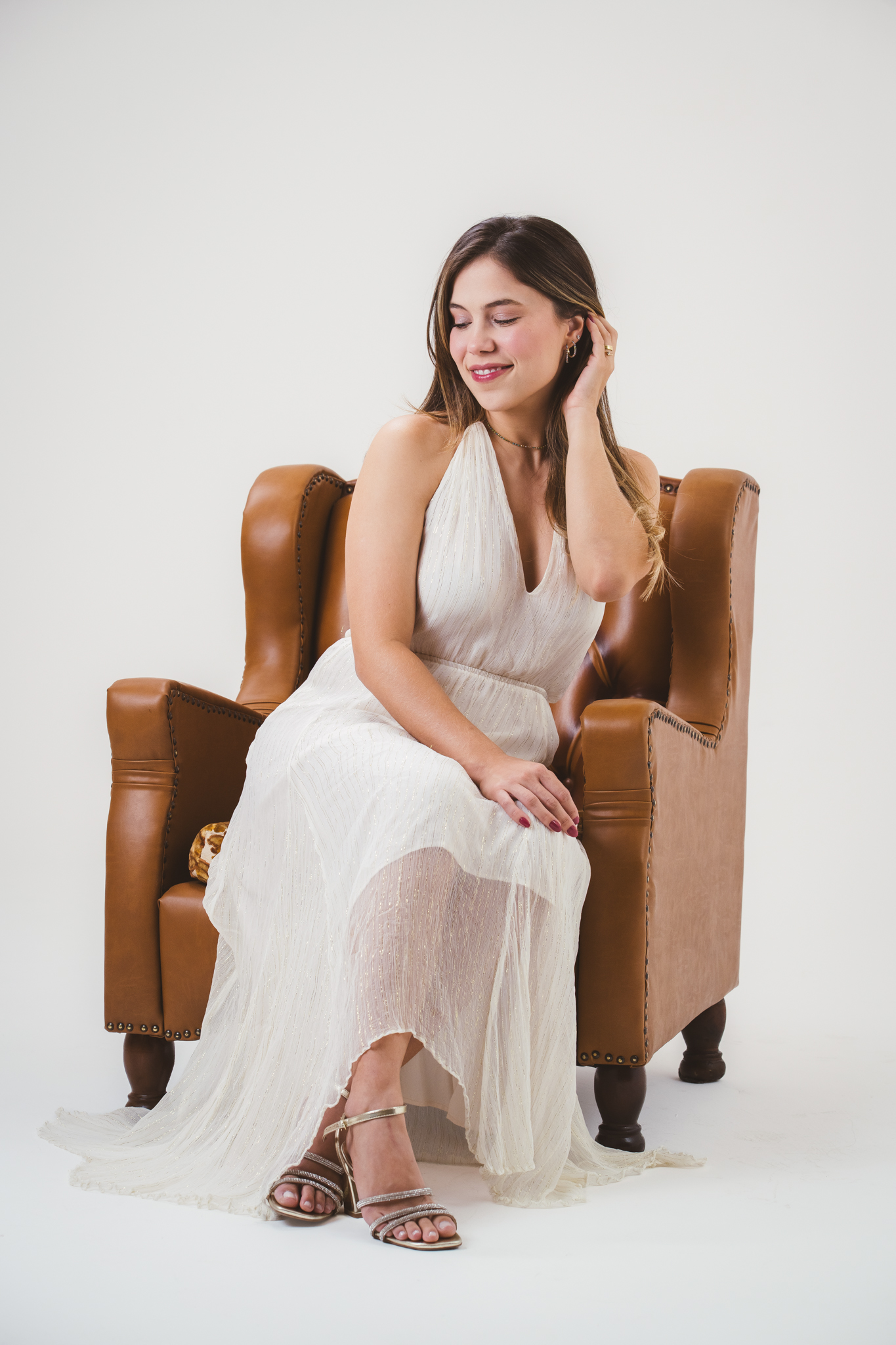 Mulher sorridente com vestido branco longo e fluido sentada em poltrona de couro marrom, sobre fundo branco.