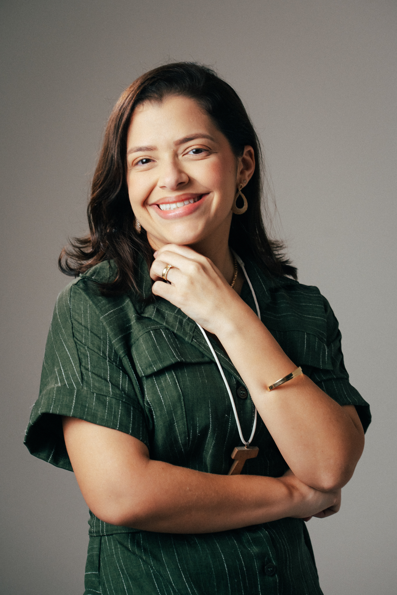 Mulher sorridente com blusa verde risca-de-giz, braços cruzados, apoiando o queixo na mão em fundo cinza.