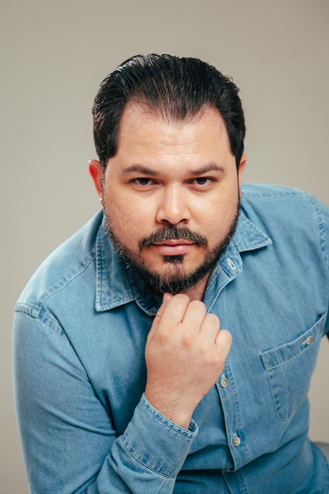 Homem barbado de camisa jeans azul, com a mão sob o queixo, olha sério para a câmera em estúdio.