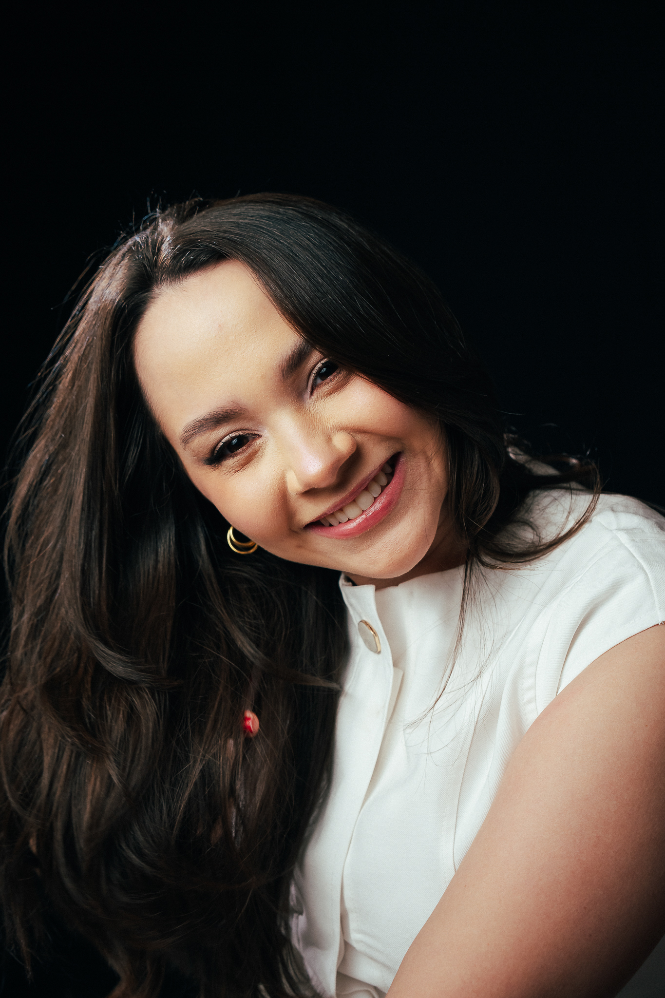 Retrato em estúdio de mulher sorridente com cabelos longos escuros e blusa branca, posando levemente inclinada contra fundo preto.
