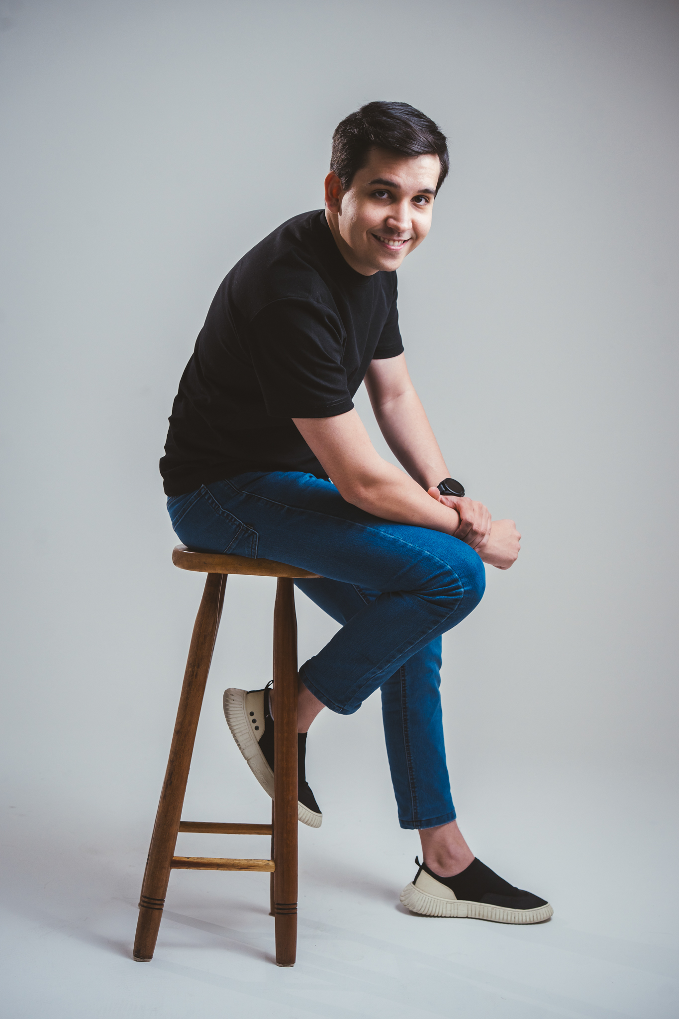 Homem sorridente de camiseta preta e calça jeans senta em banqueta de madeira contra fundo cinza claro.