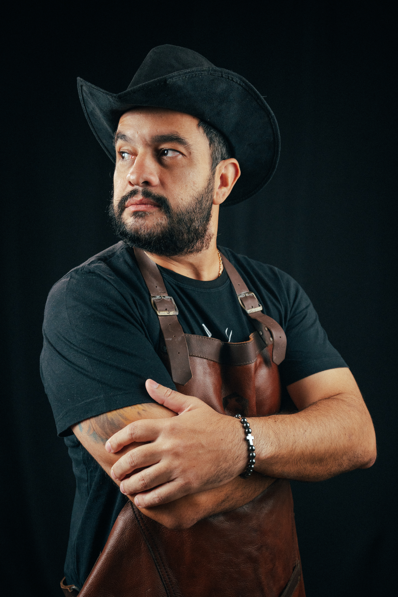 Retrato de homem barbudo, com chapéu preto, camisa escura e avental de couro marrom, olhando para o lado com os braços cruzados.