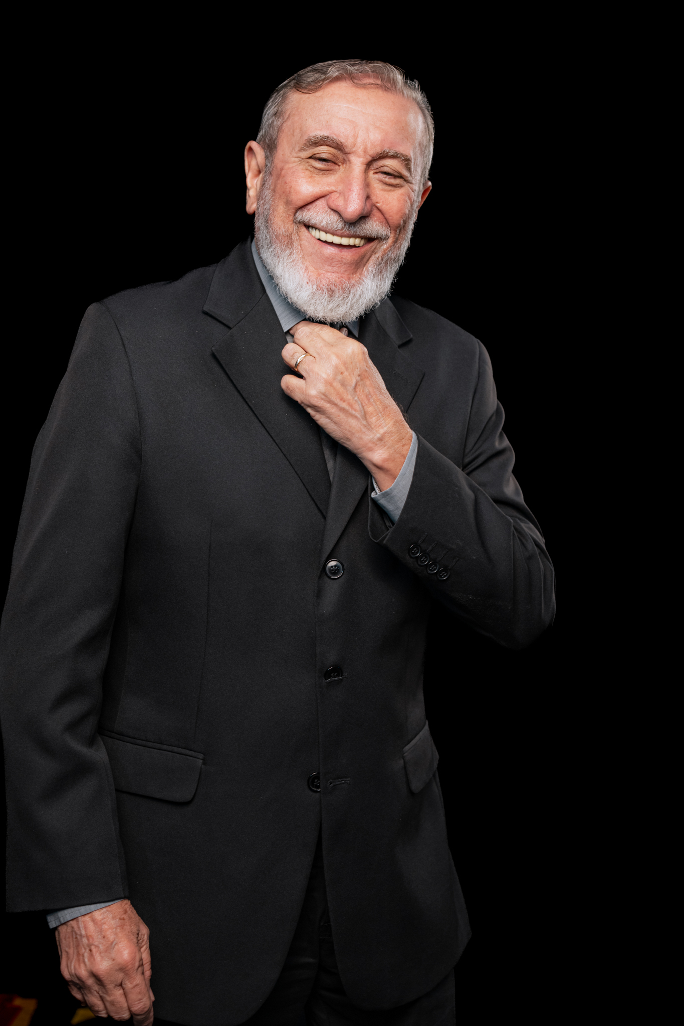 Retrato de homem madurado, sorridente, com barba branca e terno preto ajustando a gola, contra fundo escuro.