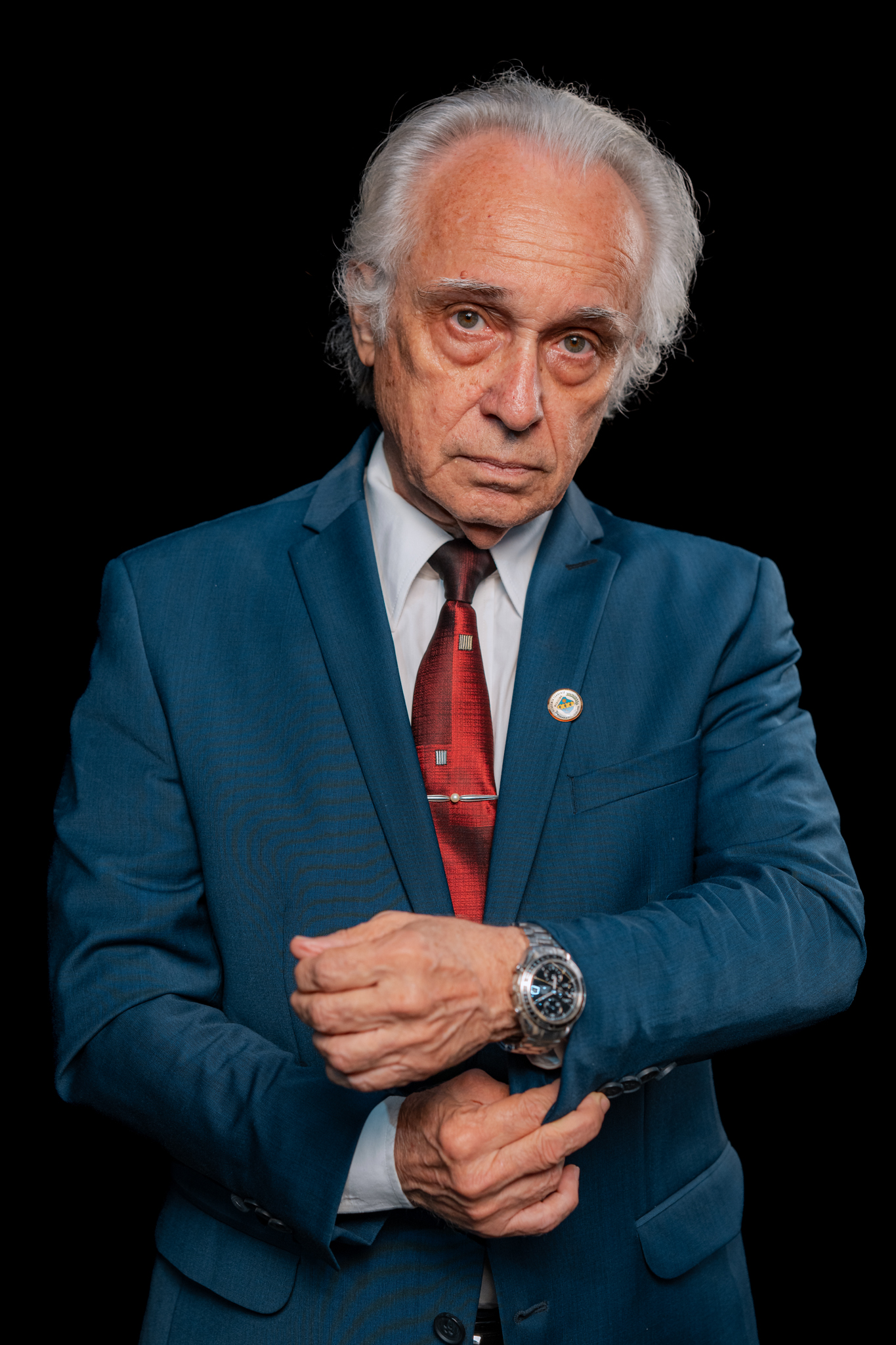 Homem idoso de cabelos brancos, terno azul, camisa branca e gravata vermelha, ajeitando o punho do paletó contra fundo preto.